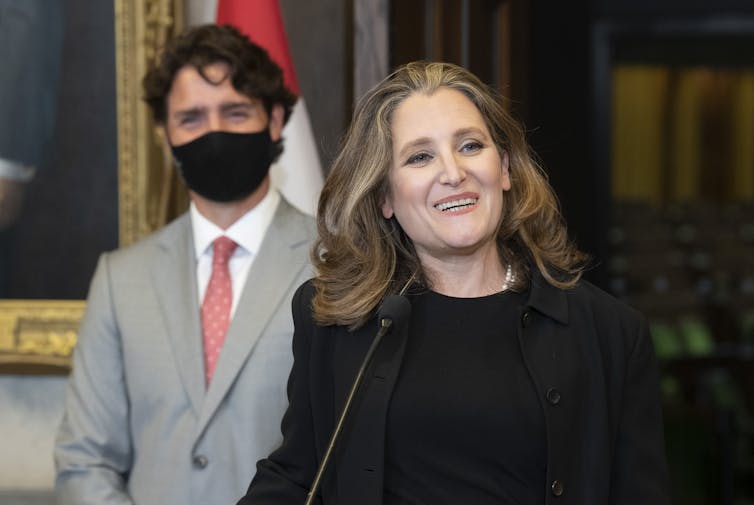Chrystia Freeland speaks at a news conference with Justin Trudeau behind her, wearing a mask.