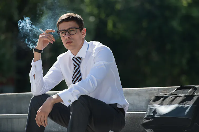 Un homme en complet cravate est en train de fumer assis sur des marches publiques.