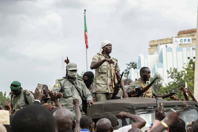 Soldiers in fatigues ride atop a jeep, with a Malian flag and large building in the background