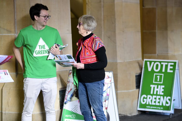 Greens candidate and volunteer, standing next to a Greens placard at a voting booth.