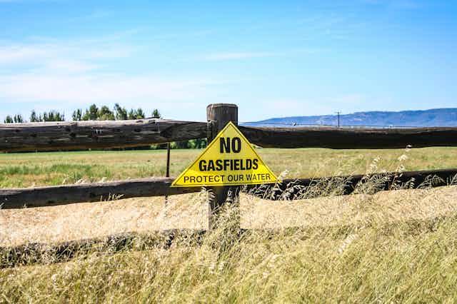 A yellow triangular sign against a wooden fence reads "NO GASFIELDS PROTECT OUR WATER"