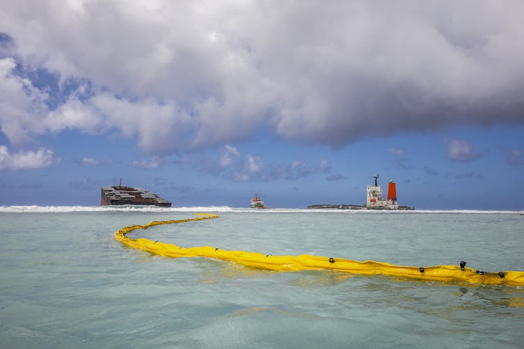 A boom rests on the ocean surface with a shipwreck in the background.