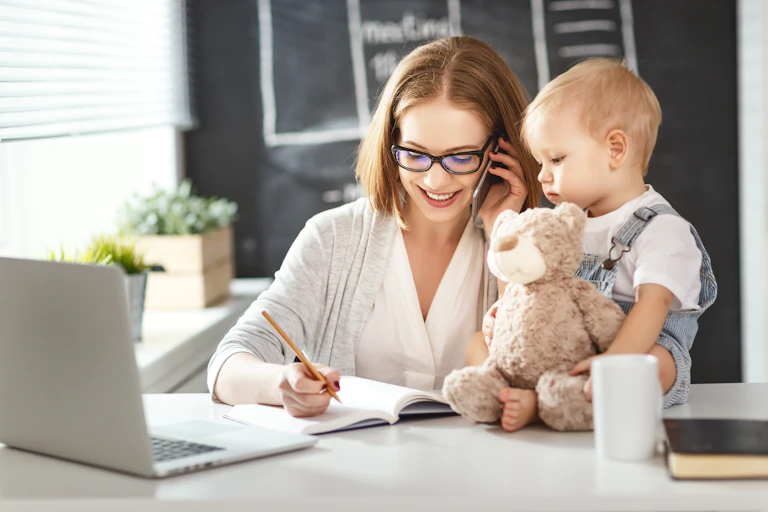 A woman working at a desk on her phone, with a baby sat next to her