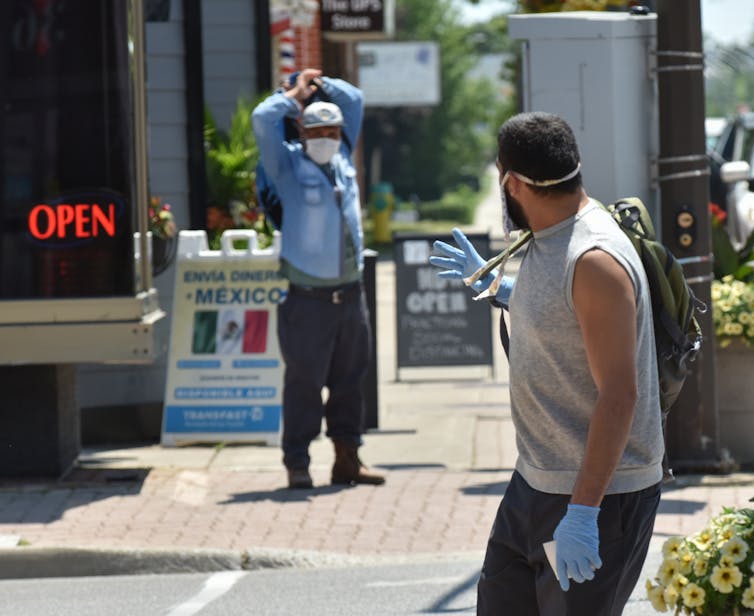 Two men wearing masks wave at one another on a street in a town.