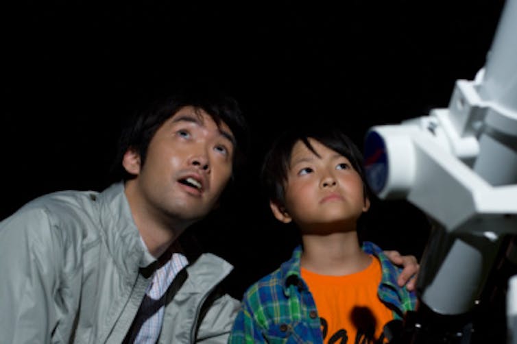 A young boy and his father view the night sky next to a telescope.