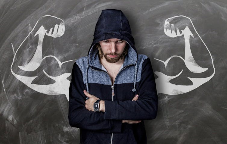 A young man in a hoodie looks pensive with his arms folded across his chest. Behind him, on a blackboard, a drawing of caricatured muscular arms lines up with his shoulders.