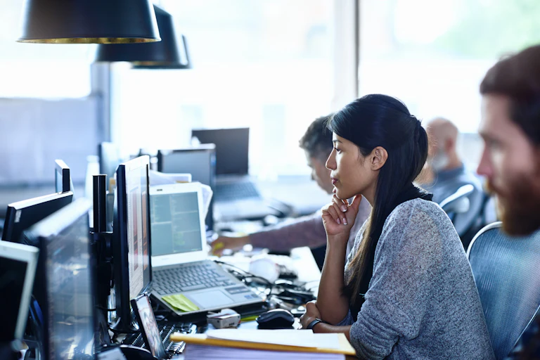 Woman and three men in an open office filled with computers. Focus is on the woman, who is staring intently at a computer screen.