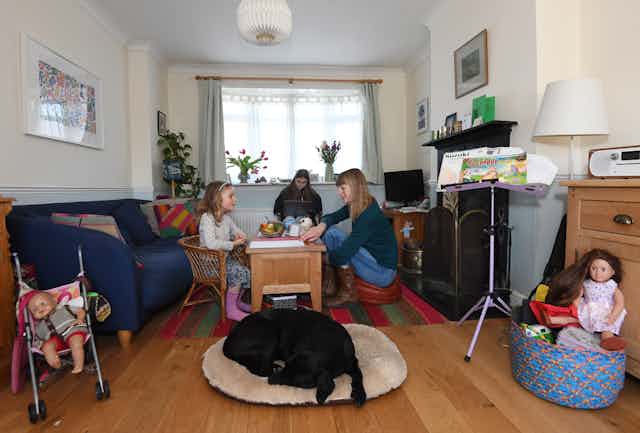 A mother teaches her children at home while a labrador sleeps by the table.