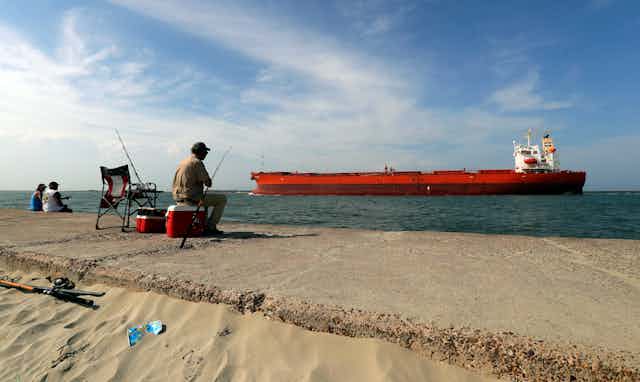 A man sitting on a cooler fishing with a red oil tanker passing by.
