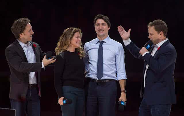 Craig Kielburger stands to the right of Sophie Gregoire-Trudeau and Justin Trudeau on a stage, with his brother Marc to their left.