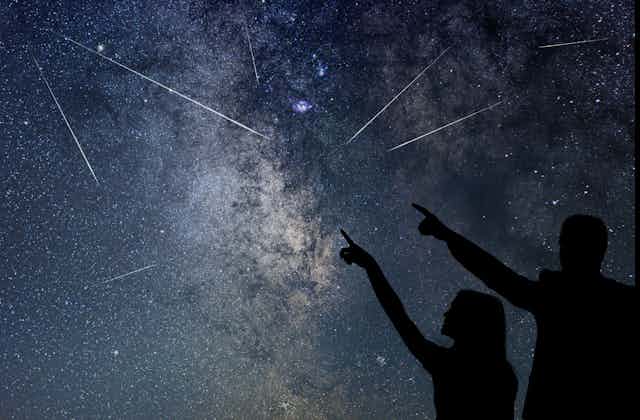 Two people point and look at shooting stars flying through the night sky. 