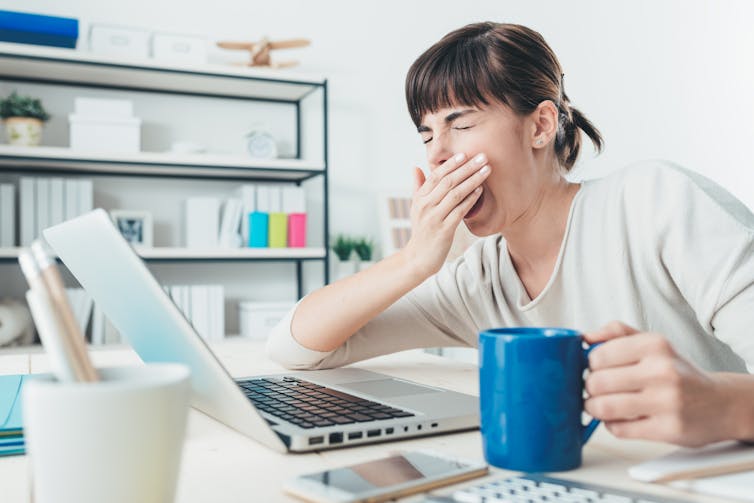 Woman holding cup of coffee yawns in front of her laptop.