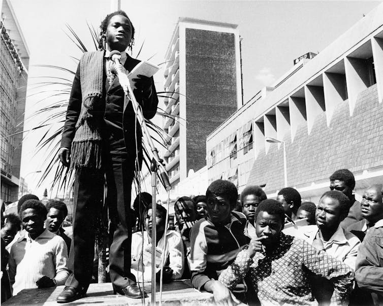 A dreadlocked man stands at a microphone, holding a notebook in an outdoor city space, crowds of people around the platform he stands on.