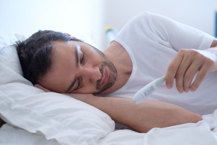 Young man lies in bed, looking at thermometer.