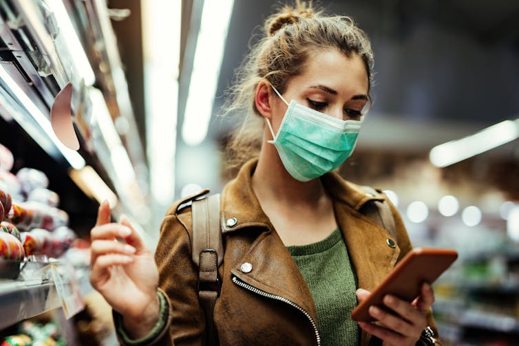 A young woman wearing a mask looks at her phone. She is in the supermarket.