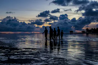 IPCC: THE THREE DECADE WAR ON TRUTH BY THE FOSSIL FUEL INDUSTRY 2 A group of people cross a shallow lagoon at dusk in the tropics.