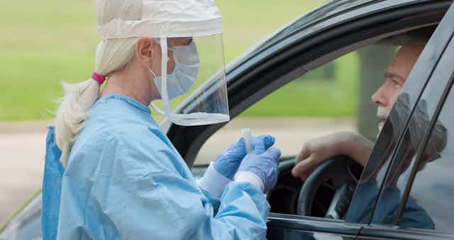 Una sanitaria protegida con mascarilla y pantalla acerca un tubo para test de saliva al conductor de un coche.