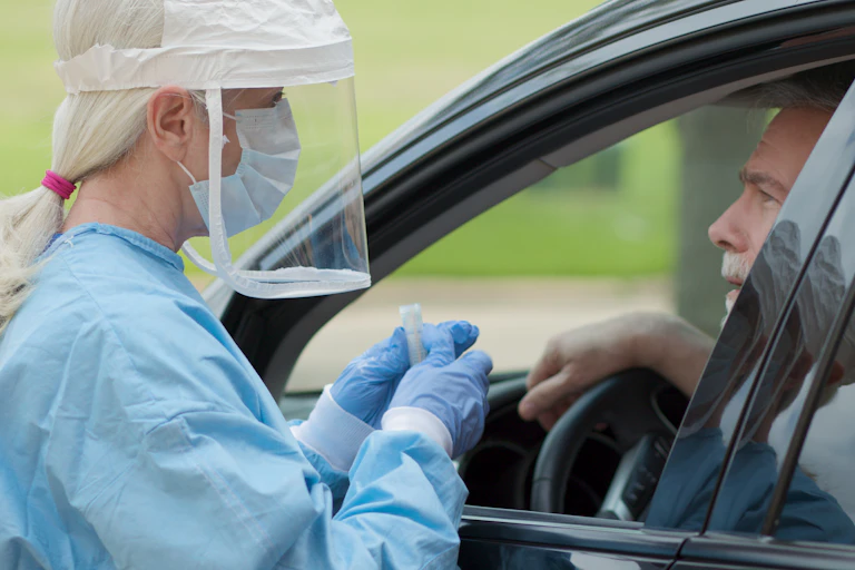 Una sanitaria protegida con mascarilla y pantalla acerca un tubo para test de saliva al conductor de un coche.