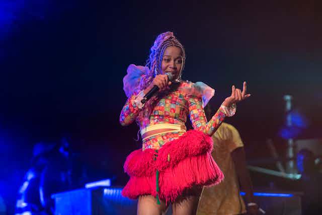 A female singer colourfully dressed in a red African skirt holds a microphone to her mouth as she performs on stage.