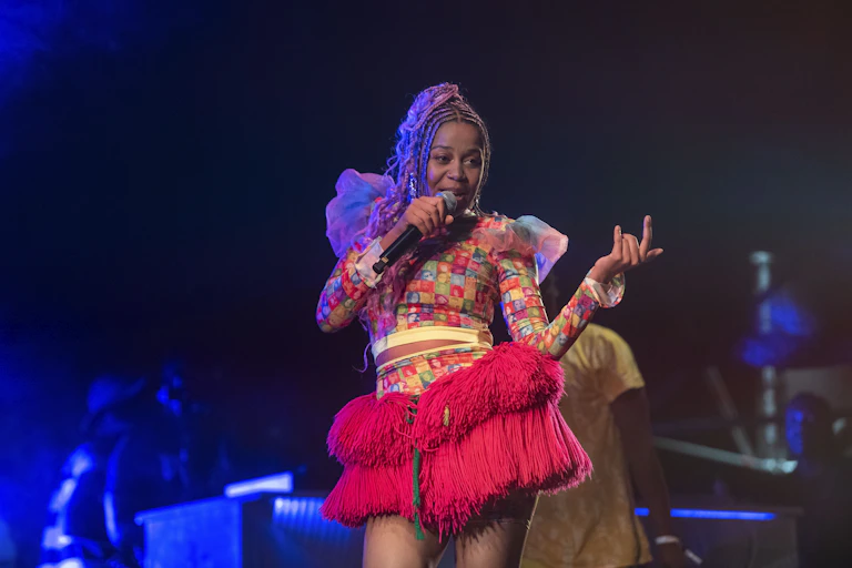 A female singer colourfully dressed in a red African skirt holds a microphone to her mouth as she performs on stage.