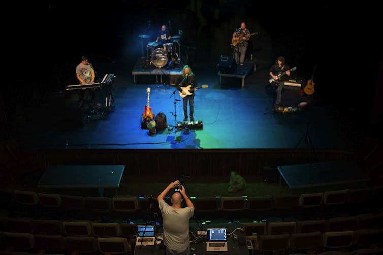 A sound engineer in an empty auditorium records a band of live musicians on a stage that is bathed in blue light.