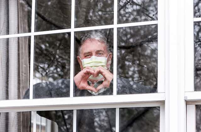 Man wearing face mask makes heart hand symbol from behind a window.