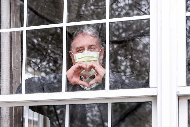 Man wearing face mask makes heart hand symbol from behind a window.