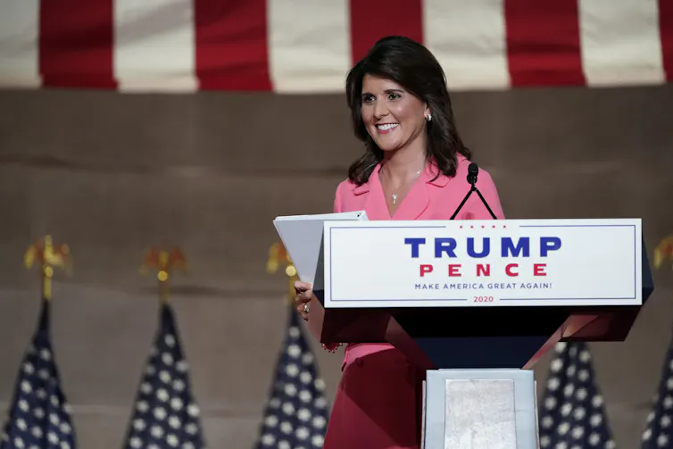 Nikki Haley, wearing a pink suit and carrying a document in her hand, smiles as she begins to walk away from a podium that reads Trump/Pence.