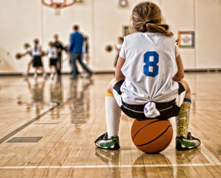 A young girl sits on a basketball while wearing a sports jersey.