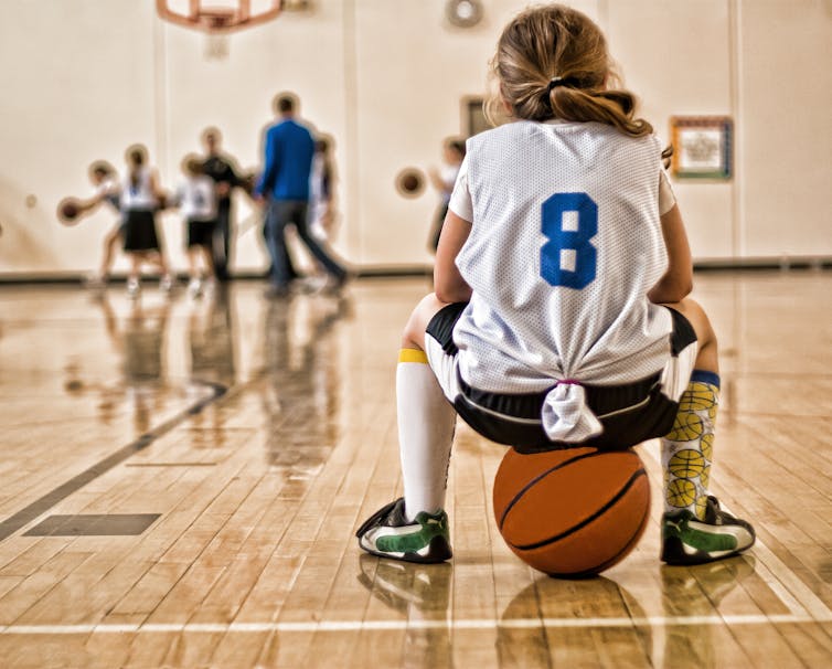 Una niña sentada sobre una pelota de baloncesto mientras usa una camiseta deportiva.