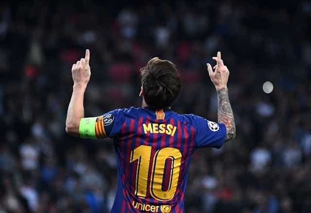 Lionel Messi de espaldas, con camiseta del Barça, señalando al cielo con los índices de las manos.