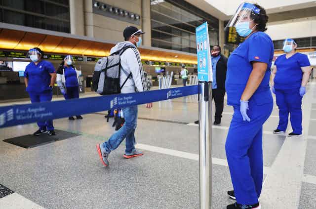 A man in jeans walks past several female airport workers dressed in scrubs.