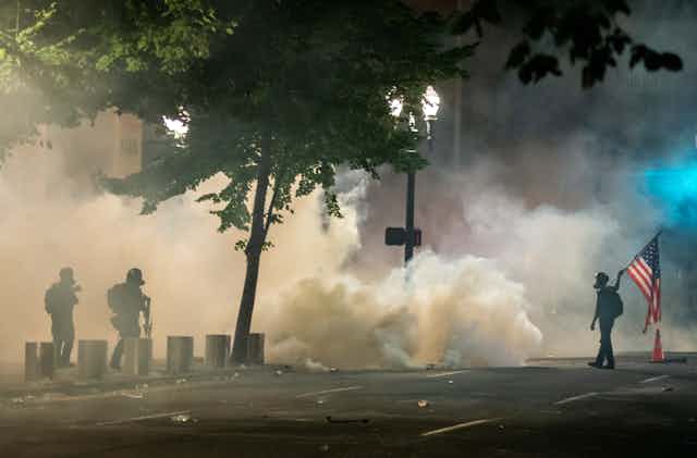 Protester carrying an American flag and two heavily armed federal agents are seen in silouhete against a backdrop of tear gas