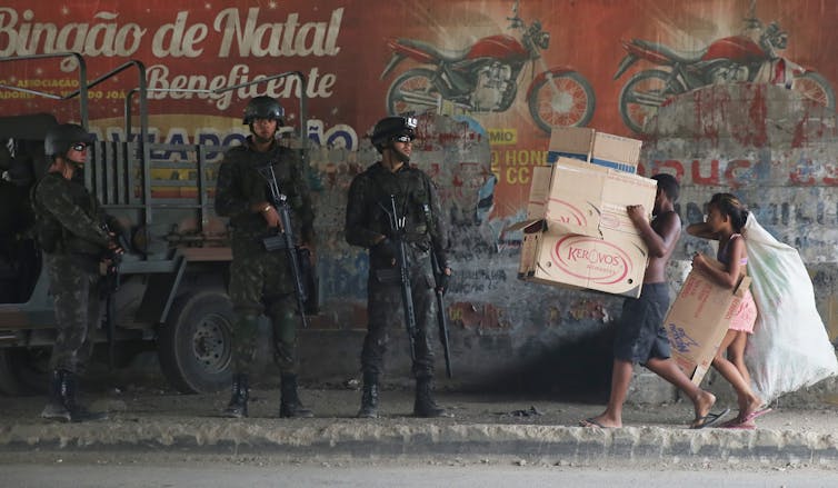 Des soldats lourdement armés regardent un petit garçon et une fille marcher en portant des boîtes
