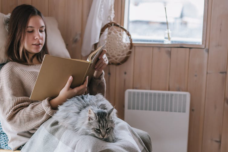 Young women in jumper and blanket with a cat sitting on her lap in front of the heater.