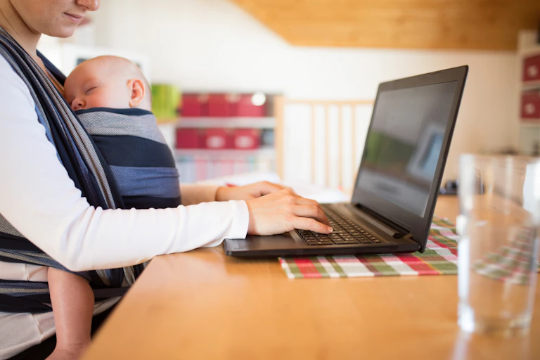 Mother with son in sling, using a laptop.