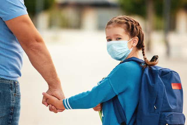 a young girl with braided hair wearing a blue cardigan and surgical mask holds her father's hand