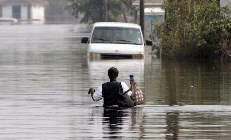 A person wades through chest-high flood water towards an abandoned car.
