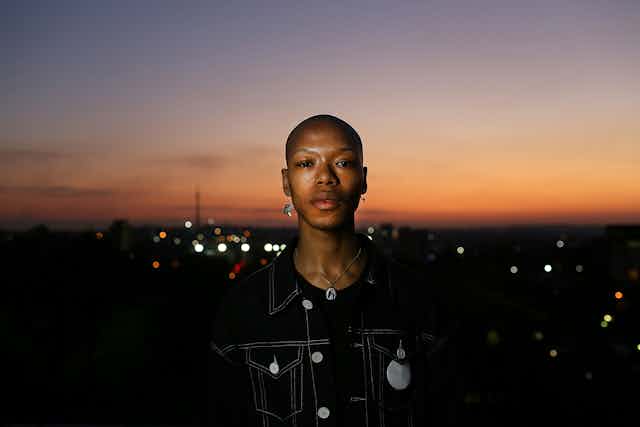 Aman with a shaved head and hanging earrings looks into camera, a cityscape of Johannesburg at sunset behind him.