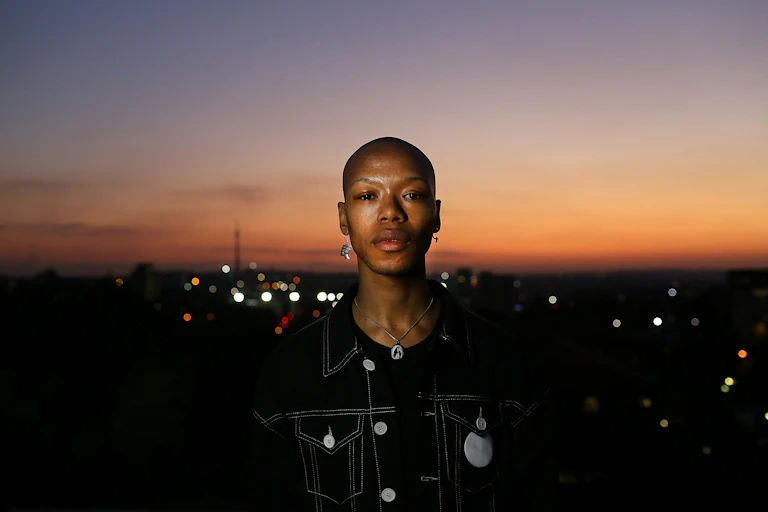 Aman with a shaved head and hanging earrings looks into camera, a cityscape of Johannesburg at sunset behind him.