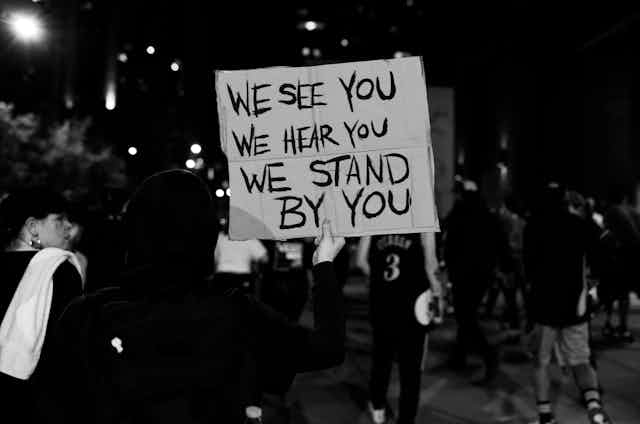 a black and white photo of a sign being held up during a protest that says "We See You. We Hear You. We Stand By You."