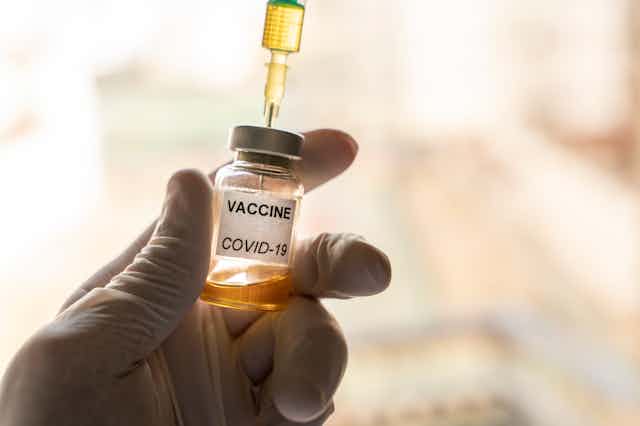 A vial marked VACCINE COVID-19 is held up by a gloved hand.