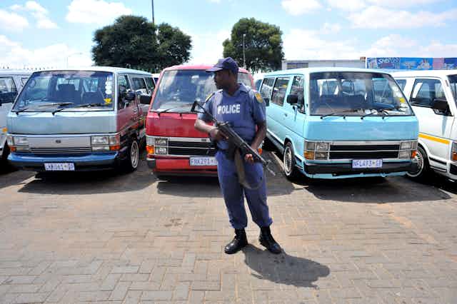 A policeman with a rifle in a parking lot guards empty minibus vehicles that form a row behind him