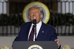 A grimacing Trump speaks into a microphone wearing a blue suit and blue and red striped tie.
