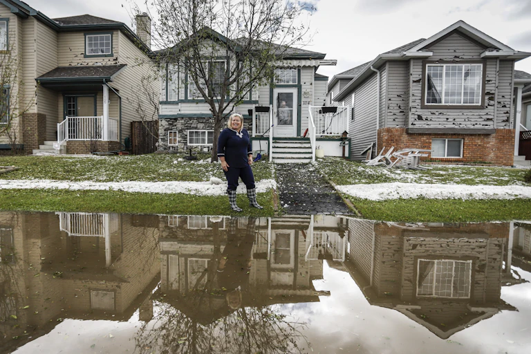 A flooded street lined with houses with damaged siding and snow on the green grass