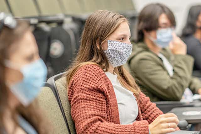 A female college student wearing a mask sits in a class next to other students with her laptop open. 