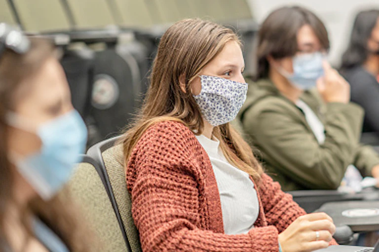 A female college student wearing a mask sits in a class next to other students with her laptop open.