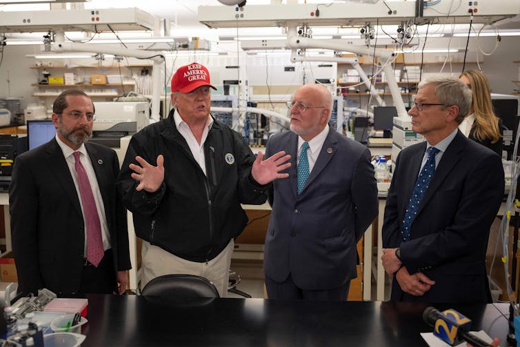 Trump, wearing khaki pants and a MAGA hat, surrounded by health officials at the CDC.