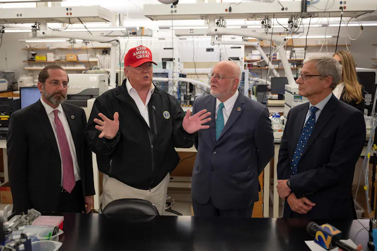 Trump, wearing khaki pants and a MAGA hat, surrounded by health officials at the CDC.
