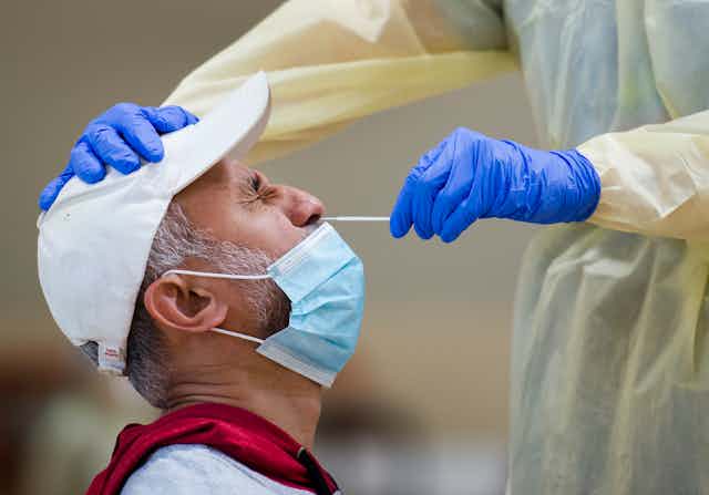 A man in profile wearing a white baseball cap and a face mask pulled down below his nose, winging as the gloved hand of a health-care worker inserts a test swab into his nose.